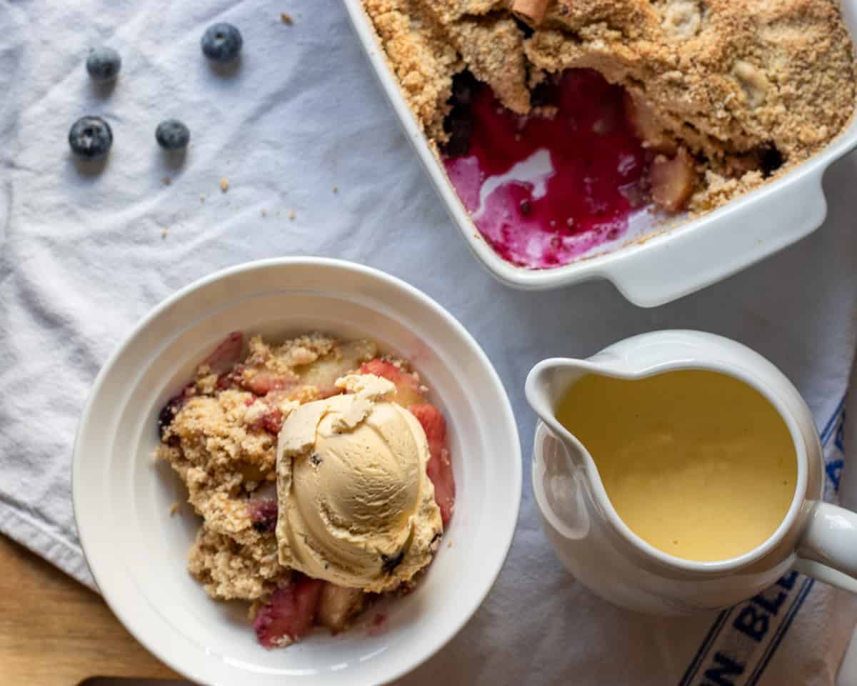 apple and blueberry crumble served with ice cream on top and custard on the side