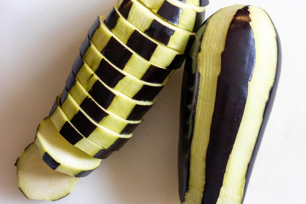 1 peeled and 1 sliced eggplant on a white cutting board.