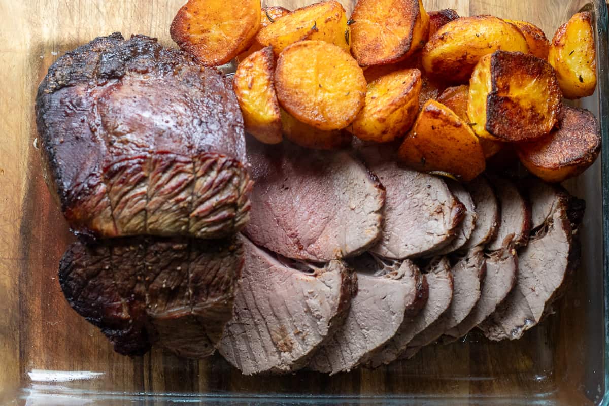 Beef sirloin tip roast is sliced and served with duck fat roast potatoes on a wooden board.