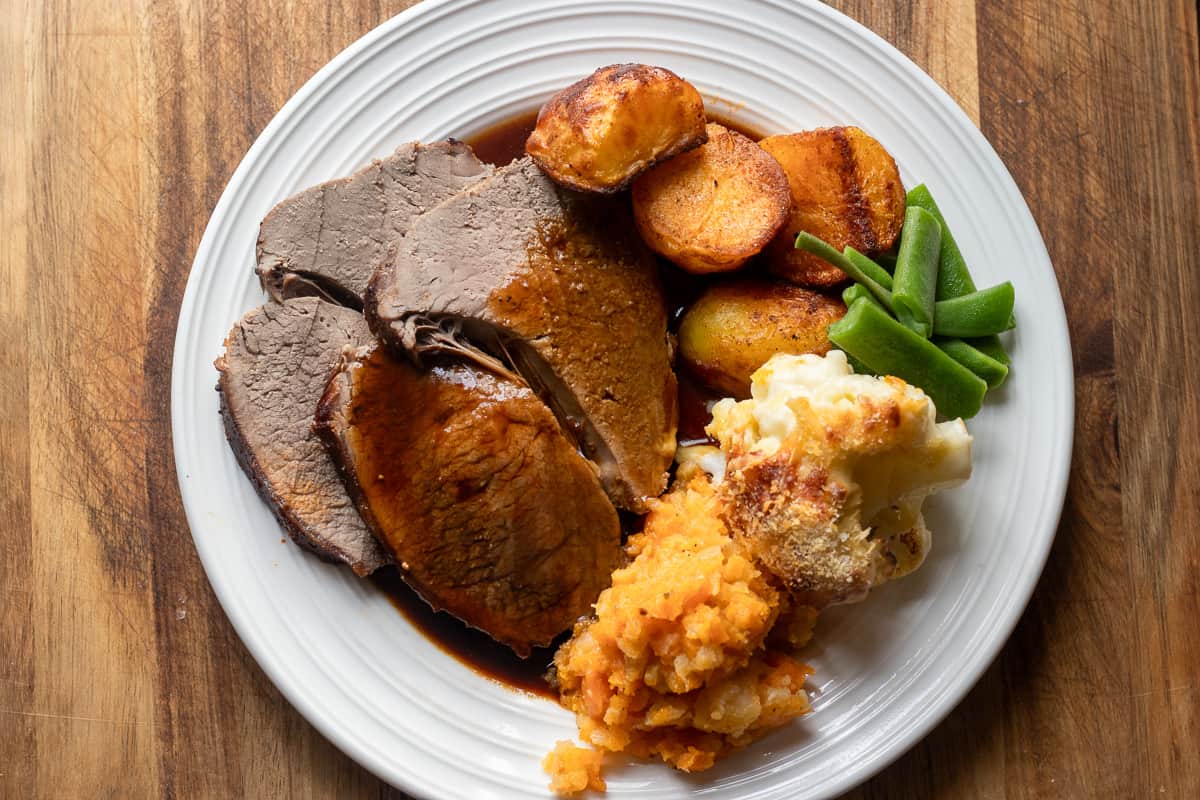The beef slices are served on a white plate with carrot and swede mash, cauliflower cheese, gravy, and roast potatoes.