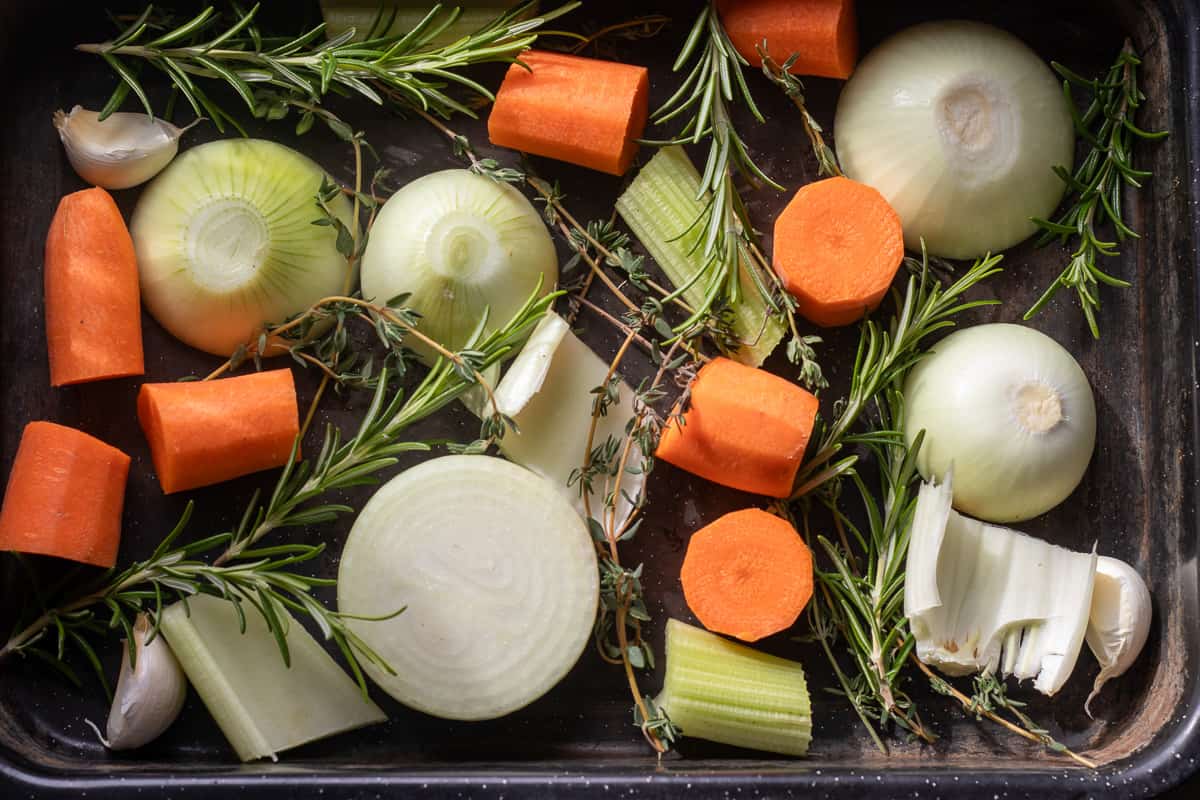 Chopped onions, carrots, celery, herbs, and garlic are layered on a baking tray.