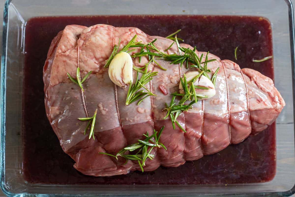 The beef is marinating in a baking dish with garlic and rosemary leaves on top.