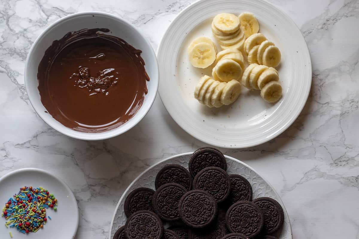 Melted chocolate, sliced bananas, Oreo cookies, and sprinkles are ready for making chocolate covered oreos.